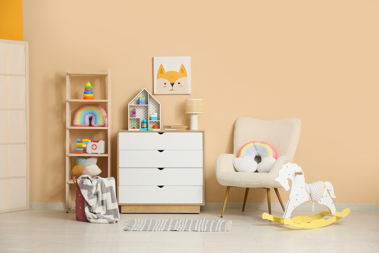 Interior of children's room with armchair, many toys in shelving unit, rocking horse and lamp on commode near beige wall