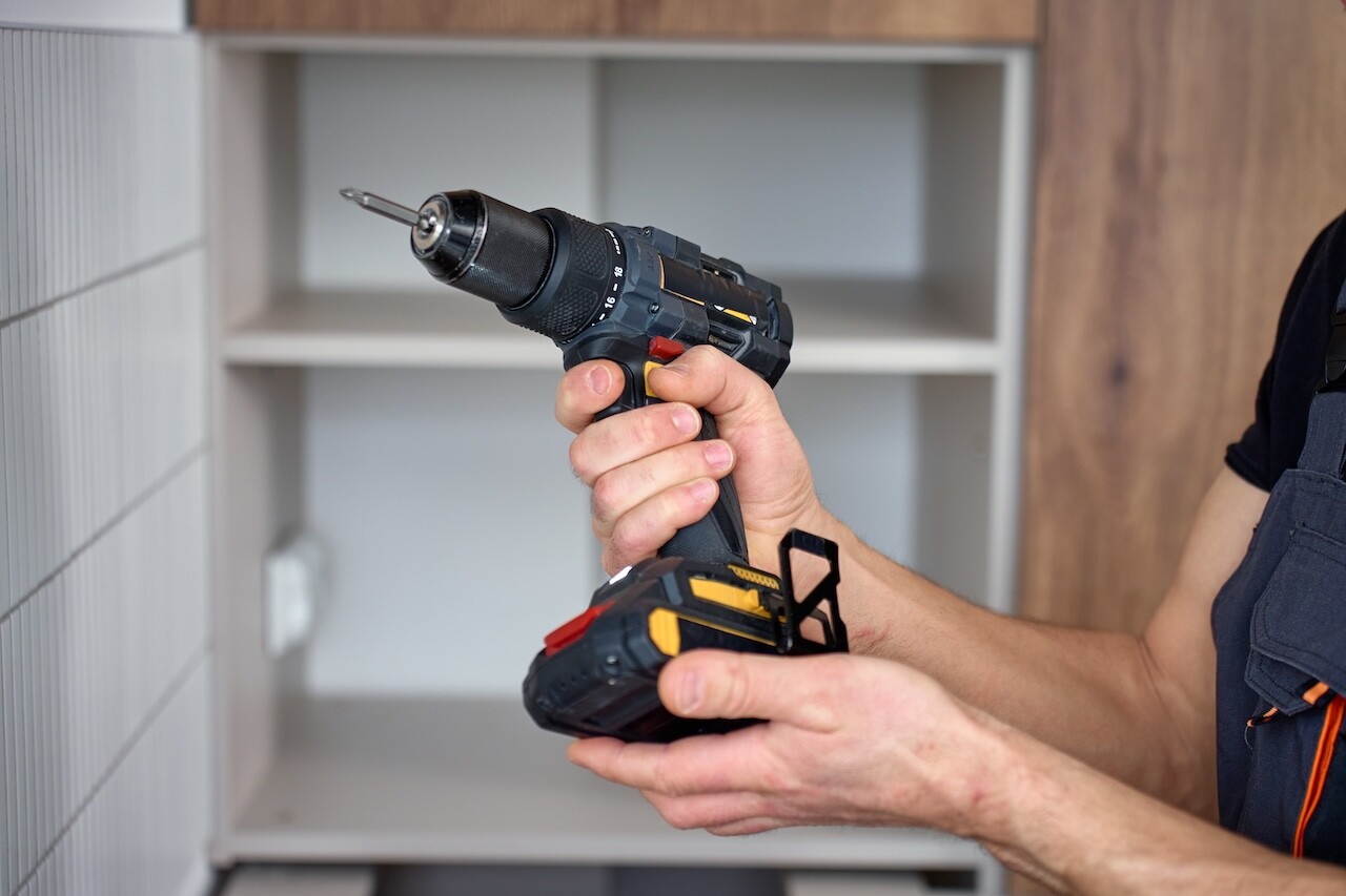 Close up of cordless drill with screwdriver bit in hand against kitchen cabinet during installation. Concept of DIY home repair and using power tools